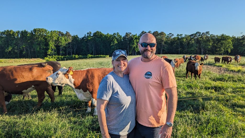 A man and woman stand smiling in front of a herd of cattle. 