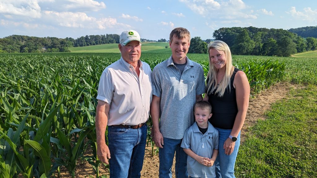 A grandfather, husband, wife and son stand in front of a corn field. 