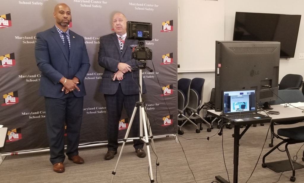 A behind the scenes shot showing the webinar setup. Dr. Tarik Harris and Michael Rudinski stand in front of a MCSS backdrop in front of a camera. 