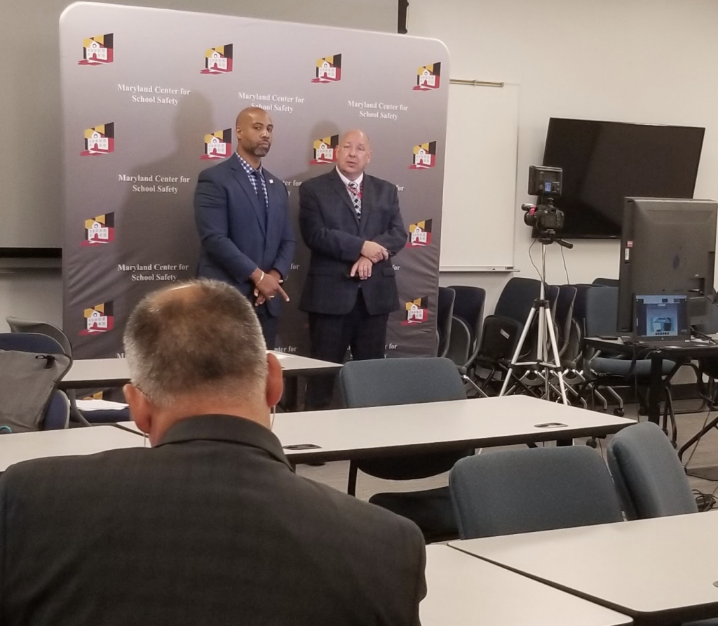 A behind the scenes shot showing the back of James Hott's head. Dr. Tarik Harris and Michael Rudinski stand in front of a MCSS backdrop in front of a camera. 