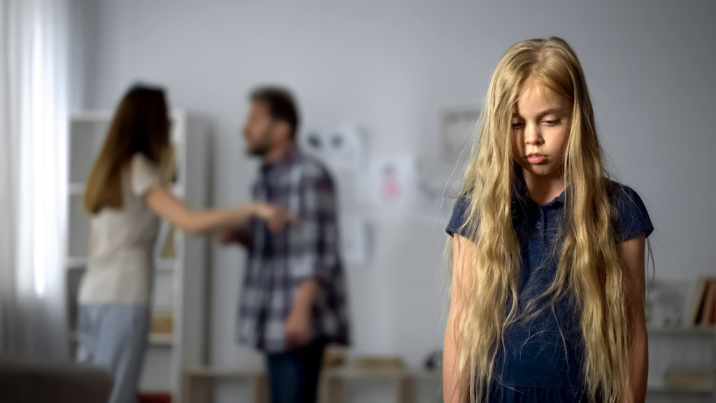 Childhood trauma and adverse childhood experiences are both addressed through Handle with Care. Image shows two adults arguing in background while child holds her head down and frowns in the foreground.