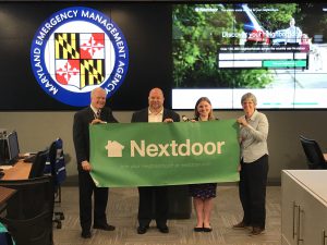 4 people are holding a long green banner that says Nextdoor on it. The Maryland Emergency Management Agency logo is displayed in the background on a screen. The website nextdoor.Com is also displayed on a different screen behind the 4 people as well.