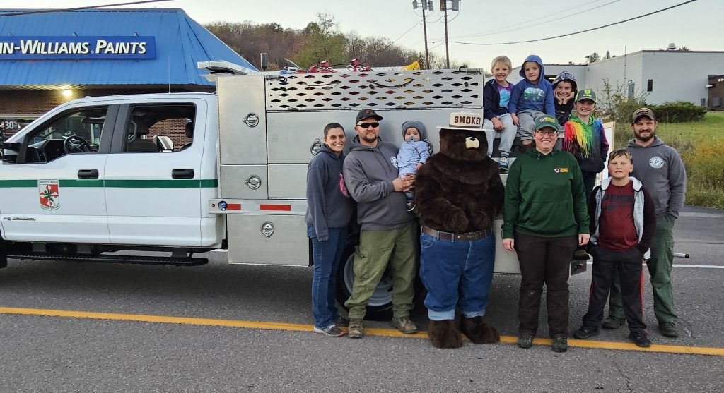 A person in a Smokey Bear costume poses with adults, children, and a woman in a Maryland Forest Service uniform in front of a forest service work vehicle