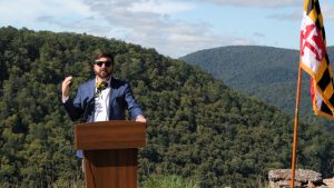 Man speaking at a podium next to the Maryland flag with a view of a valley behind him