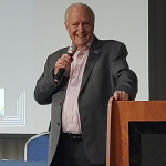 Man leans beside lectern holding a microphone with a smile on his face