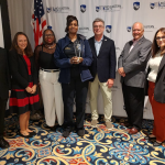 7 people are standing, posing for the photo. The woman in the middle is holding an award trophy.
