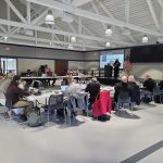 Participants sitting in a U shaped table formation are gathered listening to a presentation.